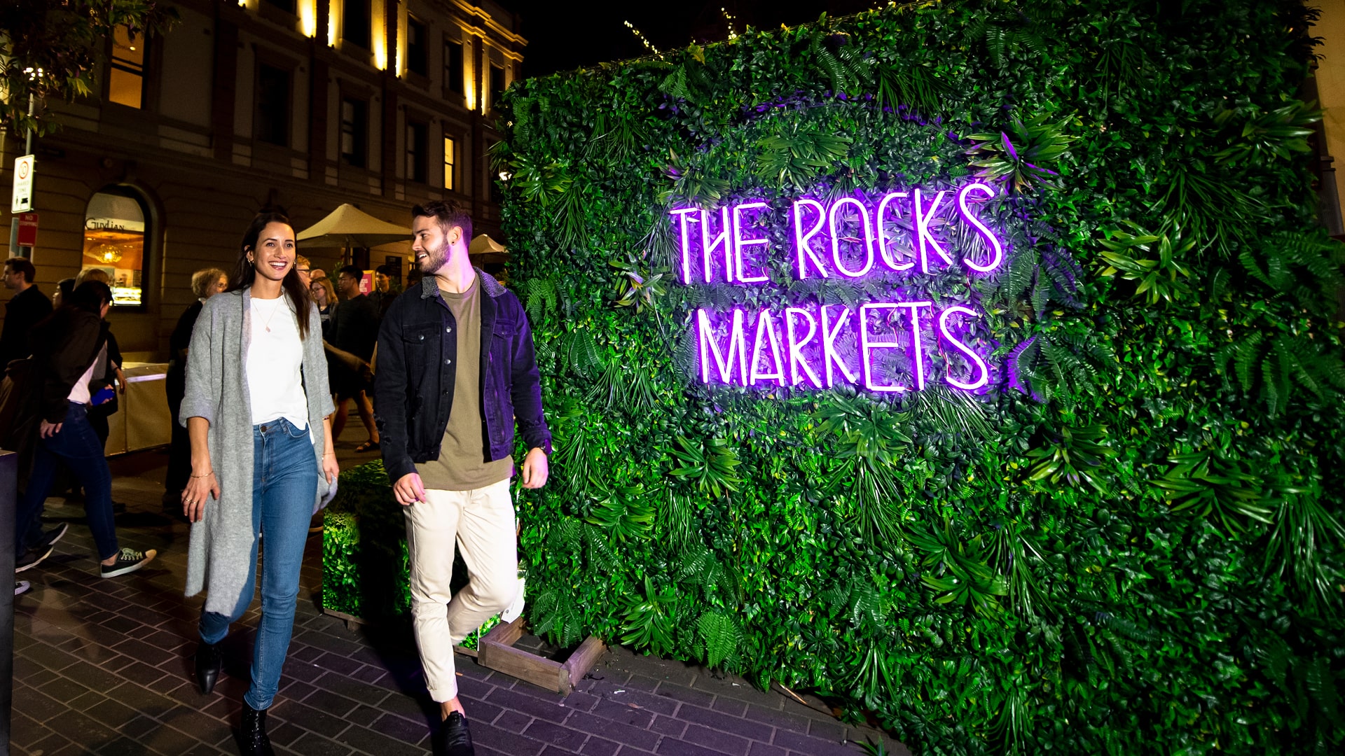 Couple walking in front of vertical garden with neon sign called the Rocks Markets